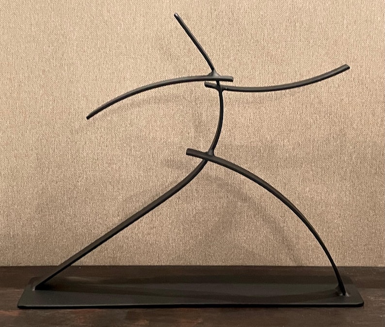 May include: A black metal sculpture depicting a stylized human figure in motion. The sculpture has a minimalist design with curved and straight lines, mounted on a rectangular base. The figure appears to be mid-stride or dancing.