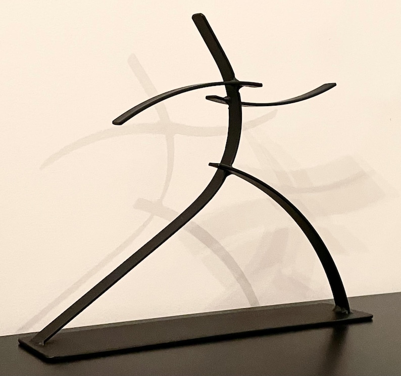 May include: A black metal sculpture depicting a stylized human figure in motion. The sculpture has a minimalist design with curved lines and a rectangular base. The figure's arms are outstretched, and its legs are bent, suggesting movement.
