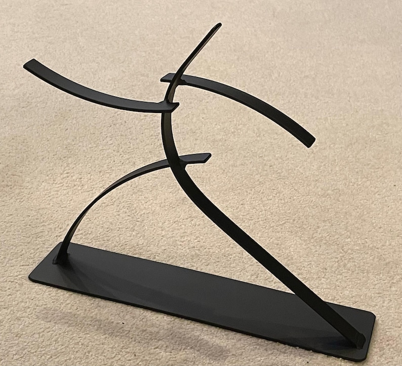 May include: A black metal sculpture depicting a stylized human figure in motion. The sculpture has a rectangular base and curved, flowing lines suggesting movement. The artwork is minimalist and modern, suitable for home decor.