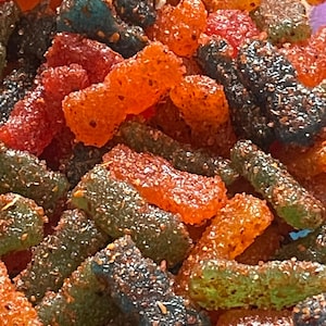 May include: A close-up shot of a pile of colorful candies. The candies are in various shapes and colors, including red, orange, green, and blue. They are coated in a granular substance, giving them a textured appearance. The candies are arranged closely together, filling the frame.