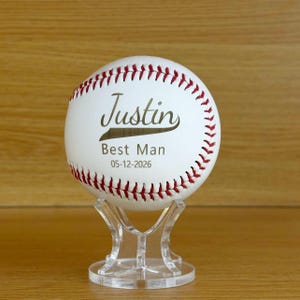 May include: A white baseball with red stitching resting on a clear acrylic stand. The ball features the printed text Justin, Best Man, and the date 05-12-2026 in a dark font. The design is centered on the front surface of the baseball.