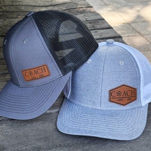 Può includere: Due cappellini da baseball con design trucker. Uno è grigio con retro in rete nera, l'altro è azzurro con retro in rete bianca. Entrambi hanno una toppa in pelle marrone con la scritta "COACH" e il nome "CHRIS - 2026".
