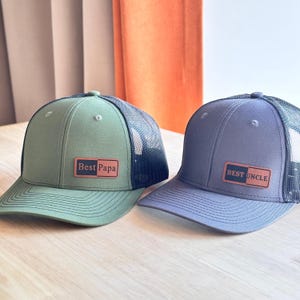May include: Two trucker caps with mesh backs. The left cap is olive green with a brown rectangular patch reading Best Papa. The right cap is slate blue with a brown rectangular patch reading BEST UNCLE. Both feature structured crowns and curved brims.