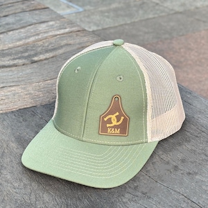 Peut inclure: Une casquette de baseball vert olive avec un filet beige à l'arrière et un écusson en similicuir marron sur le devant. L'écusson a la forme d'une étiquette d'oreille pour bétail et présente un fer à cheval doré au-dessus des lettres K&M.
