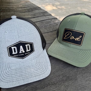 Peut inclure: Deux casquettes de baseball. L'une est grise avec un dos en filet noir et un écusson noir et blanc avec l'inscription "DAD" et "FRESH FATHER EST. 2020". L'autre est vert olive avec un dos en filet noir et un écusson avec l'inscription "Dad".