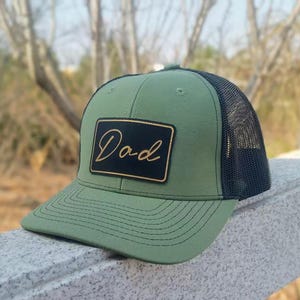 May include: An olive green trucker cap with a black mesh back and a curved peak. The cap has a rectangular patch with the word "Dad" in gold script. A fashionable accessory for casual wear.