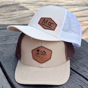May include: Two baseball caps on a wooden surface. The top cap is white with a mesh back and a brown leather patch with the letters "MR". The bottom cap is tan with a brown mesh back and a leather patch featuring a farm scene.