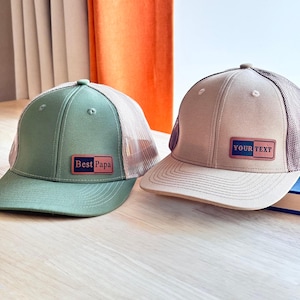 Könnte beinhalten: Zwei Trucker-Caps mit Netzrückseite. Die linke Kappe ist olivgrün und hat einen rechteckigen Aufnäher mit der Aufschrift Best Papa. Die rechte Kappe ist beige und trägt einen rechteckigen Aufnäher mit der Aufschrift YOUR TEXT.