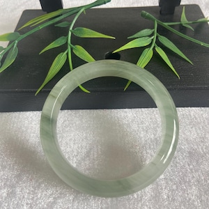 Puede incluir: Un brazalete de jade verde claro. El brazalete circular tiene una superficie lisa y pulida. El jade es translúcido, con ligeras variaciones de color. El fondo presenta elementos decorativos y follaje verde.
