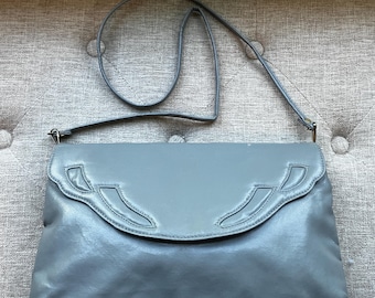 May include: A grey leather crossbody bag with a long strap. The bag has a flap closure with decorative cutouts. The bag is a soft grey colour and appears to be vintage.