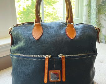 May include: A navy blue leather handbag with brown leather handles and accents. The bag features a front pocket with a rectangular logo tag and gold-toned zips. The handles are stitched with white thread.