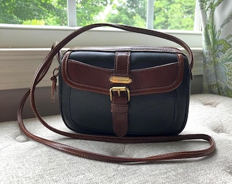 May include: A black and brown leather crossbody bag with a textured finish. The bag features a front flap with a brown leather strap, a gold-tone metal buckle, and a gold-tone metal plaque with the text Liz Claiborne centred on the front flap.