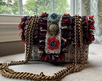 May include: A rectangular shoulder bag crafted from multi-coloured tweed fabric in shades of red, navy blue, and white. The bag features a gold-tone metal chain strap and a central vertical panel adorned with a light blue flower, a gold shell, and a red flower.