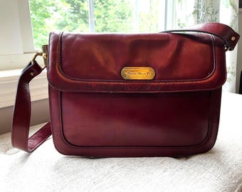 May include: A deep burgundy leather shoulder bag featuring a rectangular flap closure with a gold-toned metal plaque centred on the front. The plaque displays the engraved text Etienne Aigner and a horseshoe logo. It has an adjustable strap.