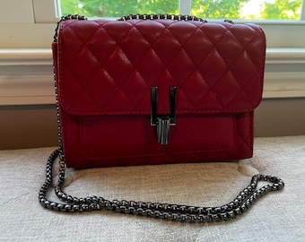 May include: A red quilted crossbody bag with a chain strap. The bag has a rectangular shape with a flap closure and a silver-tone metal clasp. The chain strap is also silver-tone. The bag is on a beige surface.