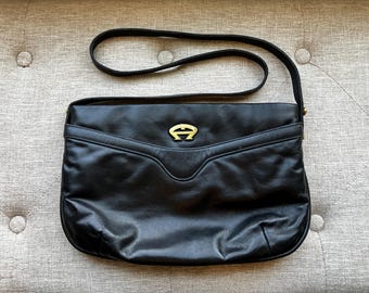 May include: Black leather shoulder bag with a long strap. The bag has a gold-coloured logo on the front and a curved design on the flap. The bag is on a neutral-coloured, button-tufted surface.