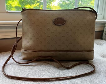 May include: Tan crossbody bag with a brown strap. The bag features a repeating triangle pattern and a brown leather patch with a logo. The bottom and trim are brown. The bag is sitting on a light-coloured surface.