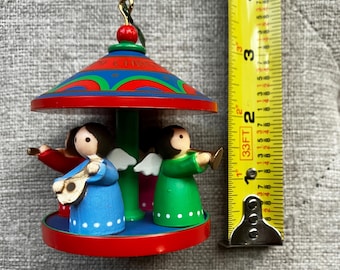 May include: A carousel-style Christmas ornament featuring three angel figures in blue, red, and green robes, each holding a musical instrument. The ornament has a red roof with blue and green patterns and is shown next to a yellow tape measure.