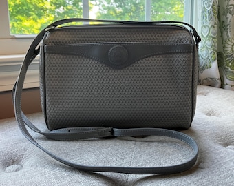 May include: Grey crossbody bag with a geometric pattern. The bag has a long, adjustable strap and a flap with a circular emblem. The bag's body has a textured, triangular design, and the trim is a darker shade of grey.