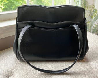 May include: A black leather handbag featuring a structured rectangular shape with a smooth finish. The bag includes two thin shoulder straps and a distinct wavy, scalloped top edge design along the main compartment and the front exterior pocket.