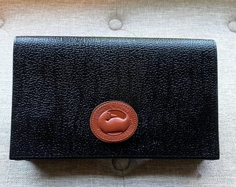 May include: Black leather wallet with a textured finish. A circular, brown leather patch featuring the "Dooney & Bourke" brand name and a duck logo is on the front. The wallet is rectangular and appears to be closed.