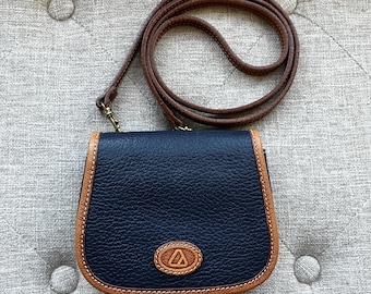 May include: A small, navy blue crossbody bag with a brown leather strap. The bag has a textured surface and is trimmed with tan leather. An oval emblem with a triangle design is centered on the front.