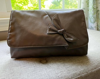 May include: A brown leather clutch bag with a bow detail on the flap. The rectangular bag has a fold-over closure and is resting on a neutral-coloured surface. The bag appears to be made of soft, supple leather.