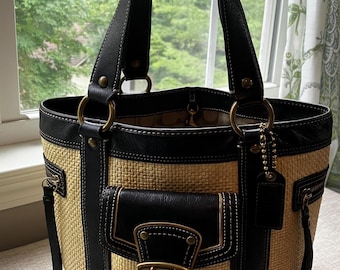 May include: A structured tote bag featuring a woven straw body in a natural tan colour, accented with black leather trim, handles, and a central front pocket with a brass-toned buckle closure. The bag includes side zipper details and a hanging leather tag.