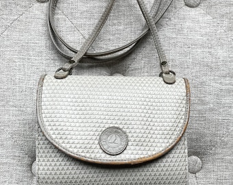 May include: A light grey crossbody bag with a textured geometric pattern and a thin shoulder strap. The front flap features a circular emblem with the text Liz Claiborne embossed in the centre. The edges of the flap show brown wear.