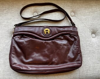 May include: A burgundy leather crossbody bag with a long strap. The bag features a gold emblem on the front and a curved design. The leather has a slightly wrinkled texture.