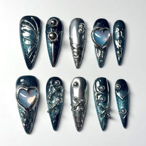 May include: A set of ten long, pointed artificial nails. The nails feature a teal glitter base with silver metallic embellishments, including spheres, heart-shaped designs, and intricate geometric patterns. The nails are arranged on a white surface.