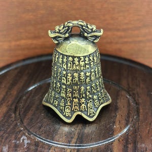 May include: A small, antique brass bell with intricate Chinese characters etched across its surface. The bell features a decorative handle with dragon-like figures. The bell is displayed on a wooden surface, showcasing its detailed craftsmanship.