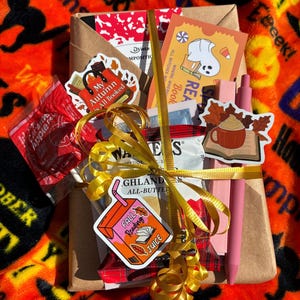 May include: A brown paper-wrapped gift set featuring a yellow ribbon, a caramel apple pop, a pack of Walkers shortbread, two pink pens, and several autumn-themed stickers including a ghost, a mug on a book, and a juice box labeled Fall Reading Juice.