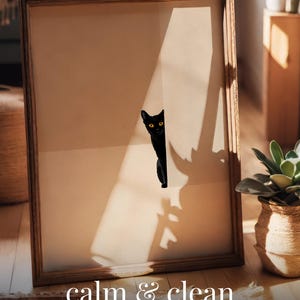 Puede incluir: Un póster enmarcado con una ilustración de un gato negro sobre un fondo beige. El póster incluye las palabras "calm & clean" y "CAT POSTER" en texto blanco. El marco es de madera.