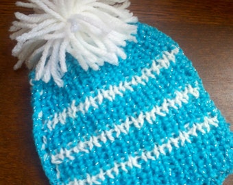 May include: A knitted blue beanie with white horizontal stripes and a large white pom-pom on top. The hat is made of a sparkly yarn, adding a touch of shimmer. This winter hat is perfect for keeping warm during cold weather.