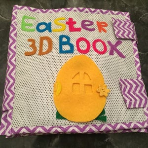 May include: A purple and white fabric book with the text "Easter 3D Book" on the front cover. The book has a yellow felt egg with a door on it.