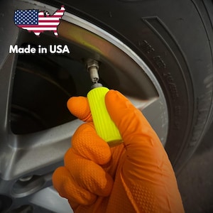 May include: A bright neon yellow tire valve cap tool held by a hand wearing an orange textured glove, positioned over a silver metal wheel rim and black rubber tire. The image includes a graphic of the United States flag and the text Made in USA.