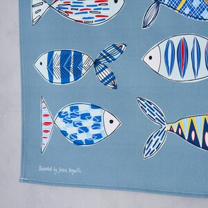 May include: A blue tea towel with a repeating pattern of colorful fish. The text "Illustrated by Jessica Hogarth" is printed in the bottom left corner.