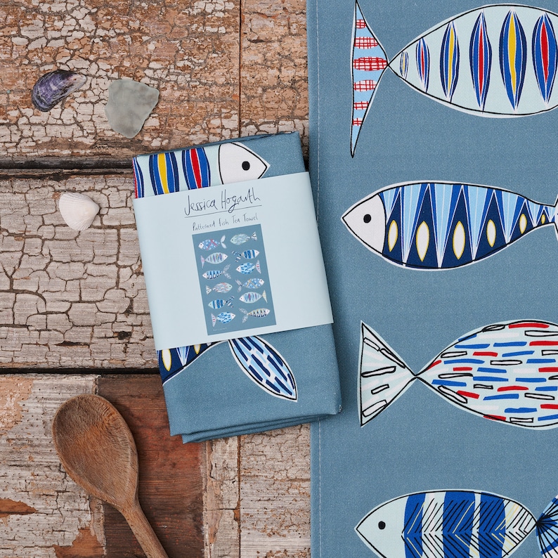 May include: A blue tea towel with a pattern of colorful fish. The towel has the text "Jessica Hogarth" and "Patterned Fish Tea Towel" printed on it.