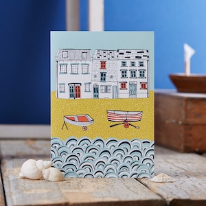 May include: A greeting card featuring a colourful illustration of houses on a beach with two small boats in front of them. The houses have red doors and windows. The boats are red and white with yellow oars. The background is a blue and white wave pattern.