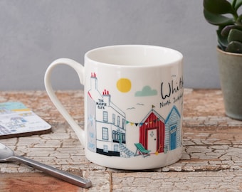 May include: White ceramic mug with a colorful illustration of a seaside town, including a cafe called "The Magpie Cafe" and beach huts. The text "Whitby, North Yorkshire" is printed on the mug.