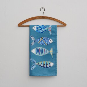 May include: A blue tea towel with a pattern of four colorful fish. The towel is hanging on a wooden hanger with the text "J. Dixon & Son, Tailors, County Durham" printed on it.