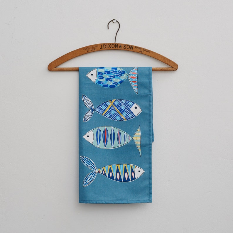 May include: A blue tea towel with a pattern of four colorful fish. The towel is hanging on a wooden hanger with the text "J. Dixon & Son, Tailors, County Durham" printed on it.