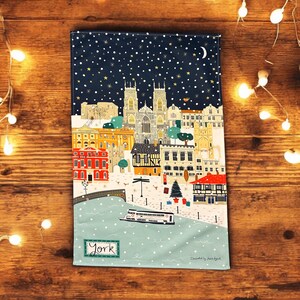 May include: A blue tea towel with a snowy cityscape of York, England. The illustration features a church, buildings, a river, and a boat. The text "York" is printed in the bottom left corner.