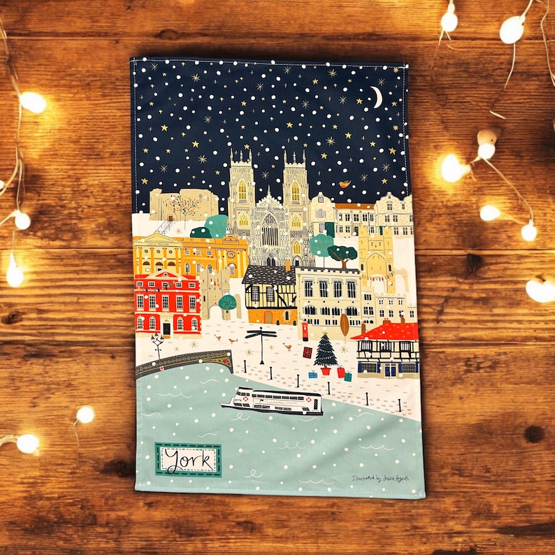 May include: A blue tea towel with a snowy cityscape of York, England. The illustration features a church, buildings, a river, and a boat. The text "York" is printed in the bottom left corner.