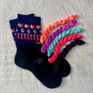 May include: A pair of dark navy blue knit socks featuring a patterned cuff with orange fish, purple crescent shapes, and pink vertical lines, accompanied by five small twisted skeins of yarn in orange, purple, turquoise, pink, and dark navy blue.