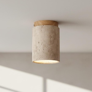 May include: A cylindrical ceiling light fixture with a light beige, textured stone body and a natural wood top. The light shines downward, illuminating the space below. The fixture is mounted on a white ceiling.