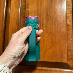 Puede incluir: Un enfriador de latas de color verde azulado con un dise&ntilde;o de lazo. El enfriador est&aacute; dise&ntilde;ado para contener una lata de bebida de tama&ntilde;o est&aacute;ndar. El lazo est&aacute; en la parte superior del enfriador y el asa es de color verde azulado s&oacute;lido.