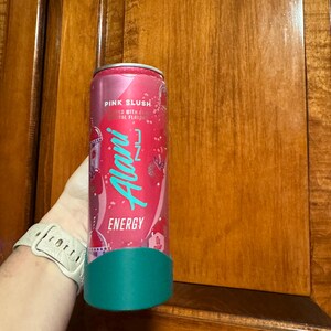 Puede incluir: Una lata de bebida energ&eacute;tica rosa con el texto "Pink Slush" y "Alani Nu Energy" en escritura turquesa. La lata tiene una base turquesa y se sostiene frente a un armario de madera.