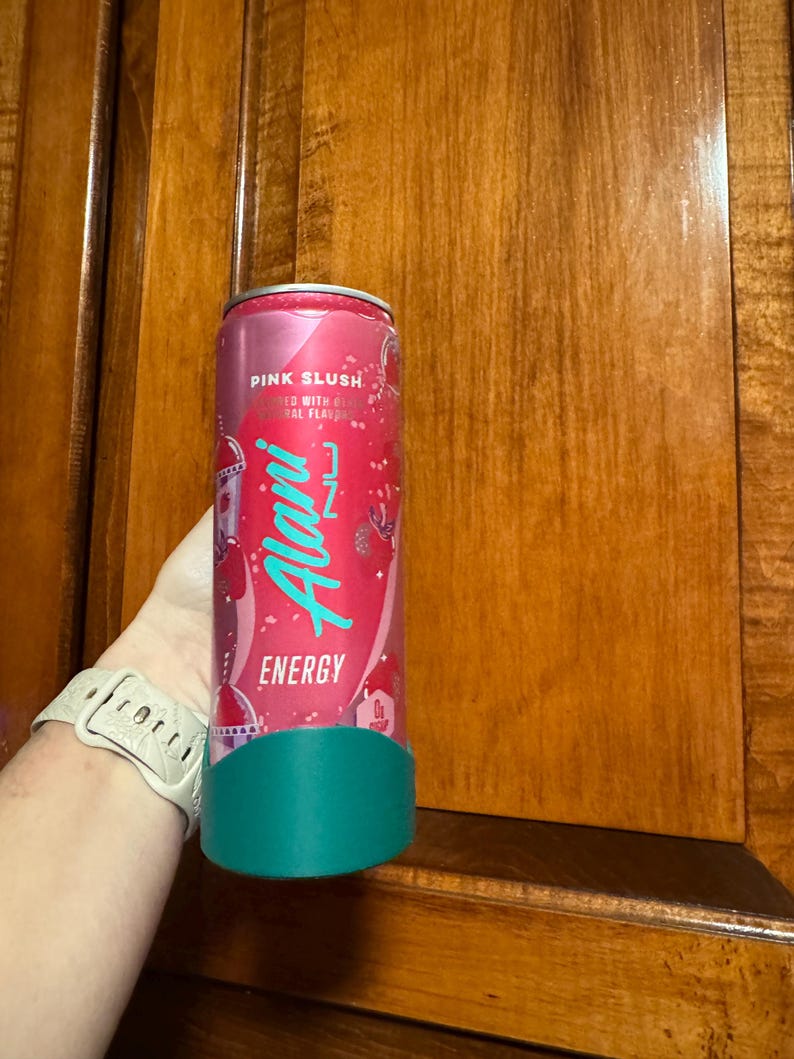 Puede incluir: Una lata de bebida energ&eacute;tica rosa con el texto "Pink Slush" y "Alani Nu Energy" en escritura turquesa. La lata tiene una base turquesa y se sostiene frente a un armario de madera.